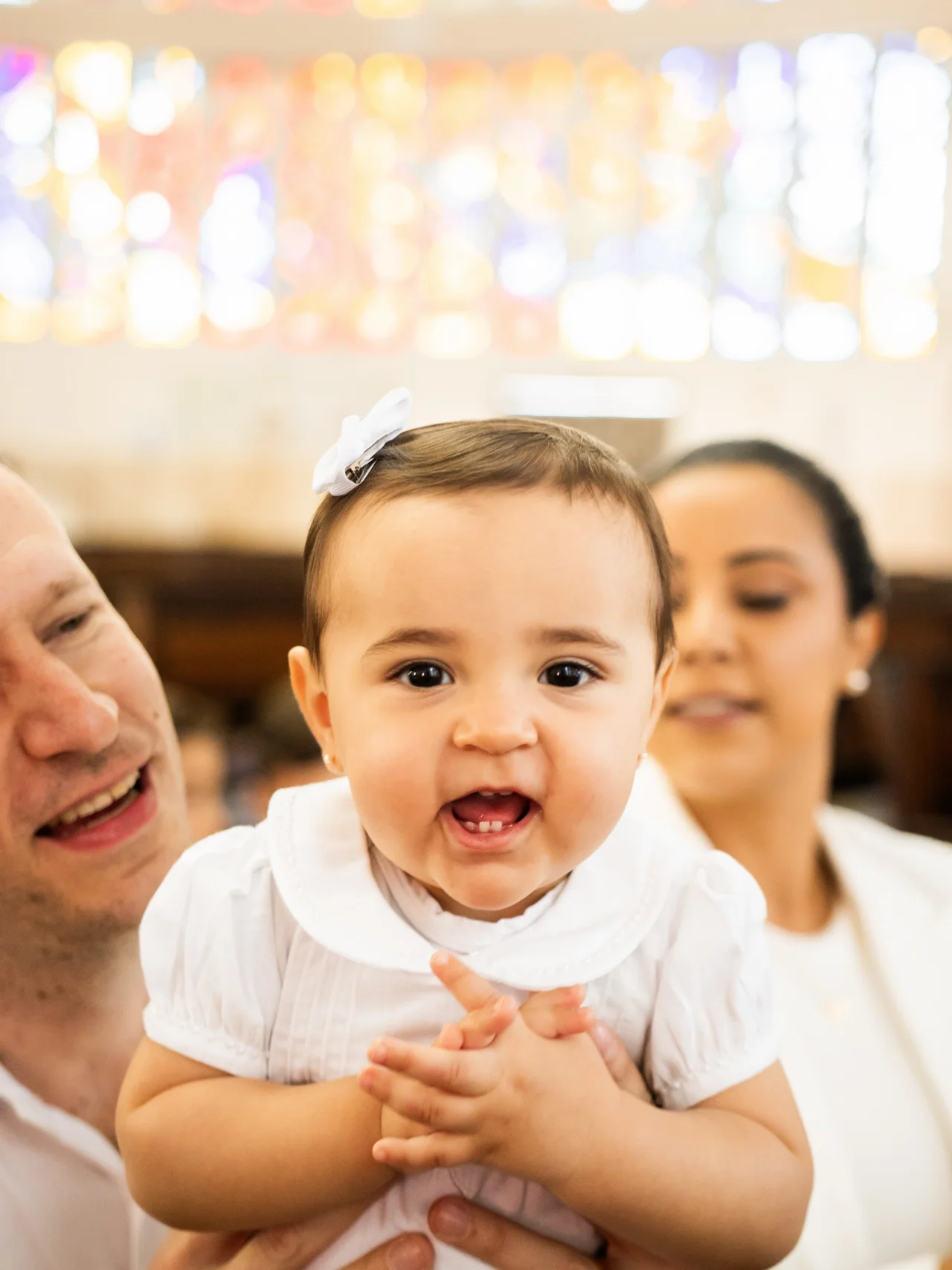 Bebê sorri entre os pais durante seu batizado.