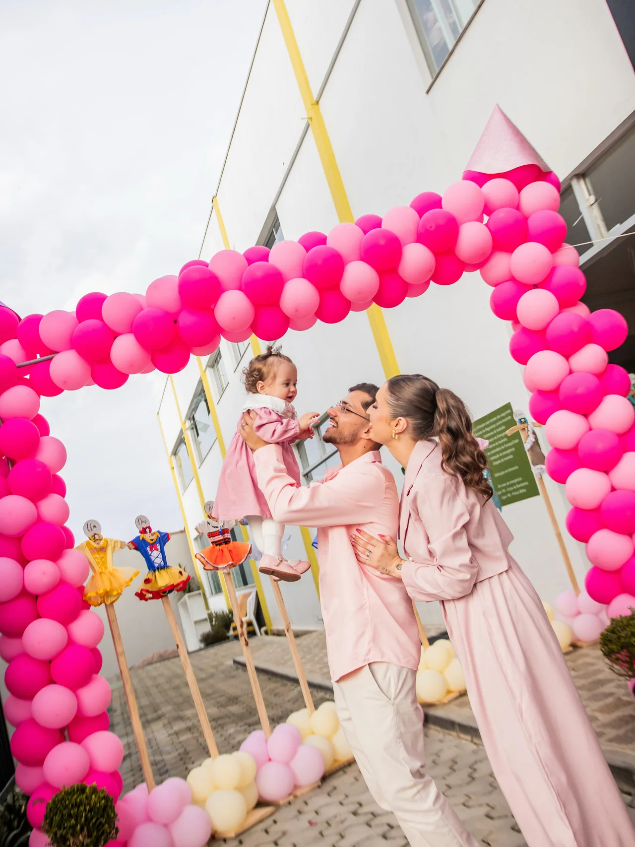 Pais erguem a filha sob um arco de balões rosa no primeiro aniversário.